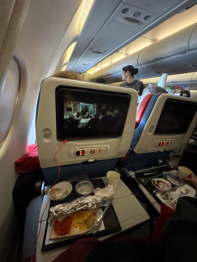 The image shows an interior view from an airplane. In the foreground, there's an in-flight entertainment screen displaying a movie, and a tray table with airline meal components, including what appears to be eggs, tomatoes, and small containers. A woman with her hair in a bun is partially visible in the background, possibly a flight attendant or another passenger. The angle indicates the photo was taken while tilting the camera. Location information points to Tambon Sa Khu, Thailand, which suggests that is the destination or departure location of the flight.