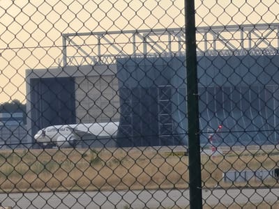 An airplane with "Lufthansa" visible on its side is parked in front of a large, dark blue building with a metal superstructure above it. The building appears to be a hangar. The scene is viewed through a chain-link fence, and dry grass and dirt are visible in the foreground. A red and white striped pole with a red light at the top is also visible in the distance. The sky is overcast and light yellow. The image was taken in Kelsterbach, Germany, near Frankfurt Airport.