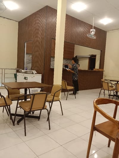 The scene depicts the interior of a dining or cafe establishment in Pandean, Indonesia. The floor is covered with white square tiles. Multiple tables are present: one square brown wooden table with four tan upholstered chairs featuring black metal frames is prominently in the foreground left, and another partial view of a similar table and chairs is visible on the far right. A light yellow support pillar stands centrally, extending from floor to ceiling.
Behind the pillar, a counter area with a brown wooden top and base spans the width of the room. Above the counter, the wall features dark brown slatted paneling, with an open serving window revealing a darker interior space. One individual, appearing to be an adult female with dark hair, stands behind the counter, facing left, holding a white object (possibly a tablet or order pad) in her hands. She is wearing a patterned short-sleeved shirt and dark trousers.
To the left of the counter, a white chest freezer is positioned next to a low metal railing. The ceiling is white and features multiple recessed square light fixtures, providing bright illumination. A white oscillating ceiling fan hangs from the ceiling above the counter area. The walls are predominantly light beige, contrasting with the dark brown paneling. No other people are visible, and the tables are unoccupied.