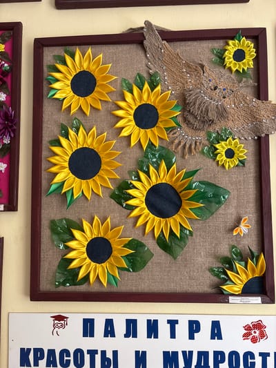 A framed artwork featuring fabric sunflowers and a jute owl hangs on a wall. The sunflowers have yellow petals and dark centers, with green leaves. The owl is made of brown jute and adorned with small blue beads for eyes and scattered sparkly embellishments. A small fabric butterfly is also present in the lower right corner of the artwork. Below the artwork, a white sign with blue text is visible, along with a small graphic of an owl wearing a graduation cap. The sign appears to be in Russian and reads "ПАЛИТРА КРАСОТЫ И МУДРОСТИ" which translates to "Palette of Beauty and Wisdom". The artwork is likely displayed in a public building or educational institution within the Vetrynsky Selsoviet, Vitebsk Region, Belarus.