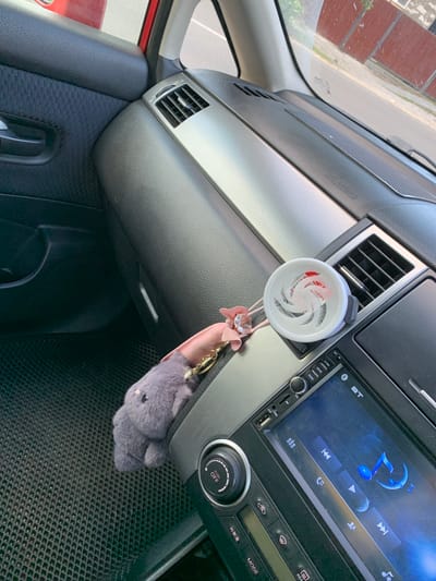 The image shows the interior of a car. In the foreground, a grey fuzzy keychain resembling an elephant is hanging from the dashboard. Next to it is a white car air freshener with a swirl design. The dashboard has a silver accent strip and an air vent. Below the air freshener is a car stereo with a touchscreen display showing music controls. To the left of the stereo is a dial with "PUSH OFF" labeled on it. The car's floor mat is visible in the lower left corner. The scene appears to be in the city of Belogorsk, Russia, as indicated by the context.