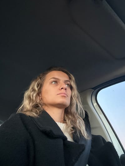 A woman with wavy blonde hair and light eyes looks upward. She wears a black coat and what appears to be a white sweater underneath. The image is taken from a low angle, showing the interior of a car, including the ceiling and sunroof. The sky is visible through the sunroof. The overall lighting is soft, creating a natural and slightly moody atmosphere.