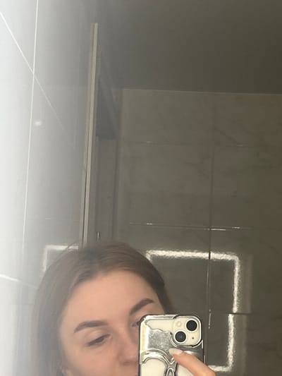 The image is a close-up selfie taken in a bathroom. A young woman is reflected in a mirror, holding a smartphone with a silver, skull-shaped case. She is looking down at her phone. The background appears to be tiled walls and a mirror with a prominent rectangular light fixture. The lighting is bright, and the overall tone of the image is slightly hazy. There is no visible indication of the location being in Nizhniye Mulli, Russia.