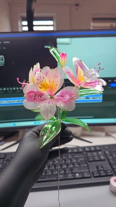 This media file shows a close-up of an artificial flower with delicate, translucent petals in shades of pink and yellow, accented with purple spots. The flower has green leaves and a wire stem. A person's hand, wearing a black glove, holds the stem, obscuring part of the flower. In the background, a computer monitor displays a light blue screen with some graphical elements, and a black keyboard is partially visible. The scene appears to be an indoor setting, possibly a workspace or craft area, given the presence of the computer equipment. The lighting suggests it is daytime, but no specific time of day can be determined. There is no visible text in the image. The overall impression is of a craft or artistic endeavor, with the focus on the handmade flower. The location context is Abinsk, Russia.