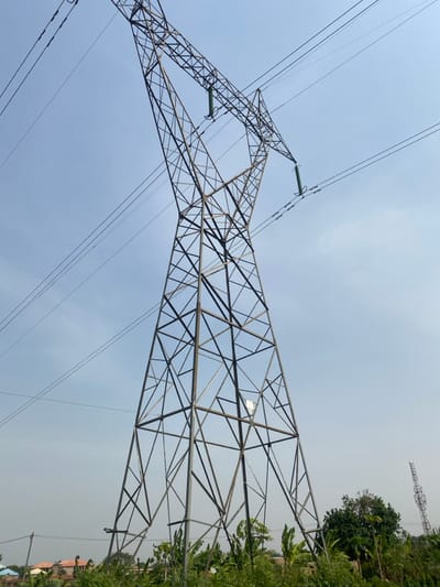 This image depicts a large, grey metallic lattice-work electricity transmission tower standing prominently against a clear, light blue sky. Multiple high-voltage power lines extend from the tower, suspended by green ceramic insulators visible in the upper section. The base of the tower is situated amidst dense green vegetation, including shrubs and what appears to be banana plants. In the mid-ground to the left, residential buildings with reddish-brown roofs and light-colored walls are partially visible, alongside a smaller utility pole. To the right in the distant background, another metallic lattice-work tower, likely a communication mast or another power pylon, is partially obscured by trees. No individuals are present. The scene is located in Fajo 2 Division Range, Nigeria, under bright daylight conditions.