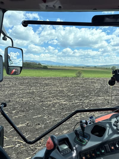 The image shows a view from inside a vehicle, possibly a tractor or heavy machinery, overlooking a field. The field is divided into two sections: one with a light green grassy area and the other dark brown and appears freshly plowed with leftover stalks. In the distance, there are hazy mountains. The sky is blue with fluffy white clouds. The interior of the vehicle is visible, including the dashboard, controls, and a side mirror reflecting the sky. The location appears to be a rural or agricultural area, with a perspective from within a vehicle suggests ongoing agricultural work in the fields. No indication of Meshtitsa, Bulgaria can be found in this image.