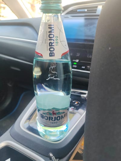 A bottle of Borjomi mineral water is placed in the cup holder of a car. The bottle is made of clear glass and is about half full of water. The label on the bottle is white with blue and red lettering. It features the brand name "BORJOMI" in large, bold letters, along with the year "1890" and the text "FROM GEORGIAN MOUNTAINS". A stylized illustration of a deer is visible on the label. The car's interior is visible in the background, including a dashboard with illuminated buttons and a screen. The overall scene suggests a person is traveling in their car and has brought the bottled water with them. The location is not specified in the image, and there is no information to suggest it is in Khorol, Russia.