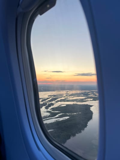 This is a photo taken from inside an airplane window, looking out at a sunset over a landscape. The view is framed by the blue interior of the airplane's cabin and the window's dark gray border. The horizon line is visible, with a gradient of colors from pale yellow and orange at the sunset to a pale blue sky above. To the right of the sunset, a dark, textured landmass is visible, likely a forest or hilly terrain, with what appears to be a body of water reflecting the sky. The image is oriented sideways, with the sunset appearing on the left side of the frame. The time of day is sunset. The overall mood is tranquil and serene, capturing a moment of natural beauty from an elevated perspective. There is no visible text in the image.