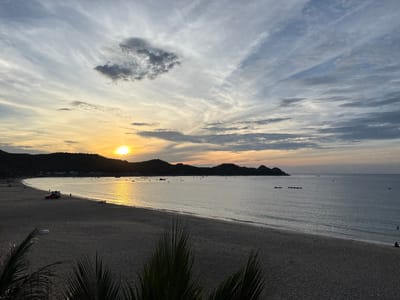 This image captures a tranquil beach scene at sunset in Song Cau, Vietnam. The sun, a bright orb of golden light, is positioned on the horizon, casting a shimmering reflection across the calm sea. The sky is a canvas of soft hues, with streaks of white and grey clouds interspersed with the warm tones of the setting sun. Silhouetted against the vibrant sky are the lush, rolling hills that frame the crescent-shaped bay.
The beach itself is a broad expanse of sand, dotted with a few sparse palm fronds in the foreground. A red vehicle with an attached cargo area is parked on the sand, near the water's edge, with a few figures standing around it. Further down the beach, other individuals can be seen strolling or sitting, enjoying the serene atmosphere. Several small boats are anchored or drifting on the water, adding to the peaceful ambiance. The waves gently lap at the shore, creating a soft, rhythmic sound that complements the visual tranquility of the scene. The overall impression is one of natural beauty and peaceful solitude, a perfect moment captured at the end of the day.