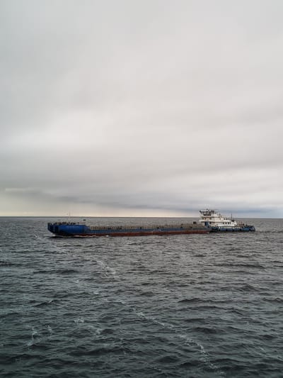 The image shows a long, flat-topped barge being pushed by a tugboat on a body of water, likely a river or lake. The sky is overcast and gray. The barge is blue with a reddish-brown hull, and it appears to be empty. The tugboat is white with blue trim and has a multi-level superstructure. The water is choppy with small waves. This is likely near Prislon', Russia, as identified.