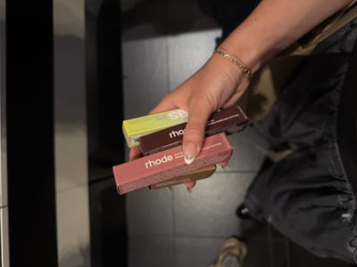 Here is a forensic description of the image:

A person is holding three small boxes of Rhode lip care products. The top box is yellow and labeled "Revealer." The middle box is brown, labeled "LIP TINT, A LEVRES TEINTE AUX PEPTIDES." The bottom box is pink, labeled "rhode PEPTIDE LIP TREATMENT." The person is wearing a gold bracelet. The background includes a black and white striped pattern and gray tile flooring. A black bag is slung over the person's shoulder. The scene appears to be indoors, possibly in a store. Marina del Rey is not explicitly evident in the image.