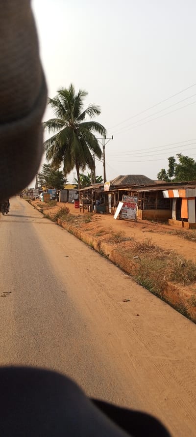 The image displays a sunlit outdoor scene along a road in what appears to be a semi-urban or rural setting, consistent with Ketu Adieowe, Nigeria. The left side features a paved road with a dirt and gravel shoulder, separated by a concrete curb. A motorcycle with a rider is visible moving down the road in the distance. A large, blurry, dark object occupies the lower left foreground, suggesting it's part of the camera operator or vehicle.

On the right, a series of simple, one-story structures, likely roadside stalls or shops, line the reddish, dusty ground. These buildings have corrugated iron roofs and appear constructed from various basic materials. A tall palm tree stands prominently behind the initial stalls, and multiple utility poles with power lines extend across the clear to hazy sky. A few individuals can be seen near these stalls, indicating daily activity and commerce.

A white sign with red and black text is clearly visible in front of one stall, displaying "PREMIUM" at the top and "POS" (likely Point of Sale) in large letters below it, followed by "AVAILABLE HERE" and other smaller text. The ground is characterized by dry, reddish soil and patches of sparse, dry grass. The lighting suggests it is either late morning or early afternoon, with soft shadows falling across the scene. The overall impression is one of everyday life and local commerce in a warm, dry climate.