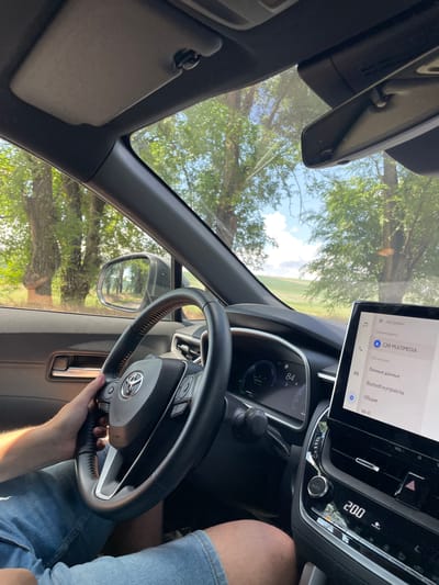 The image captures the interior of a car, looking towards the right side of the driver's seat. A person's left hand is visible, holding the steering wheel, which features a Toyota logo. The dashboard displays a digital screen showing "CAR MULTIMEDIA" and various menu options in Russian. To the left of the steering wheel, a smaller digital display shows "84 D". The car is in motion, indicated by the view of trees and a rural landscape through the windshield and side mirror. The environment outside the car appears to be a sunny day with green trees and a blue sky. The image was taken from inside the vehicle, focusing on the driver's perspective and the car's interior. The location is identified as Coșernița, Moldova, suggesting the car is traveling through this area.