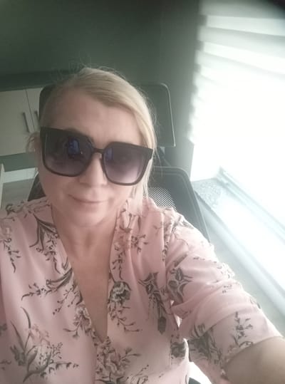 The image is a close-up, selfie-style photograph of a fair-skinned woman with blonde hair and fair skin. She is wearing large, dark-rimmed sunglasses and a light pink blouse with a delicate floral print in shades of grey and white. The blouse has a V-neck and slightly voluminous sleeves. She is seated in what appears to be an office chair, as indicated by the dark mesh material visible behind her.

The background is dimly lit and shows a hint of a dark green wall on the left and a bright, diffused light source coming from a window with horizontal blinds on the right. The light suggests it might be daytime, possibly late morning or afternoon, with the sun shining through the blinds. In the far left background, part of a cabinet with white doors is visible.

The woman is looking directly at the camera with a slight, closed-mouth smile, conveying a sense of gentle pleasantness or self-assurance. The overall mood is casual and relaxed, as if she is taking a quick break or a moment for a personal photo. The context provided places the scene in Ankara, Turkey. There is no visible text in the image.