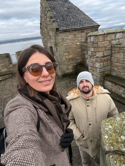 A selfie image depicts two individuals on the stone battlements of Blackness Castle in Blackness, United Kingdom, overlooking a body of water under an overcast sky. In the foreground, a female with dark hair wears amber-tinted smart glasses, a brown and cream patterned tweed coat, a brown and black striped scarf, and a black glove on her right hand. She smiles directly at the camera. Slightly behind her and to the right, a male wears a light grey beanie hat, a beige faux fur-lined winter jacket, and has facial hair; his hands are in his pockets, and his expression is neutral. Behind them, a stone building with a steeply pitched dark slate roof is visible, along with distant land across the Firth of Forth. Lichen growth appears on the top surface of a stone wall in the lower right foreground.