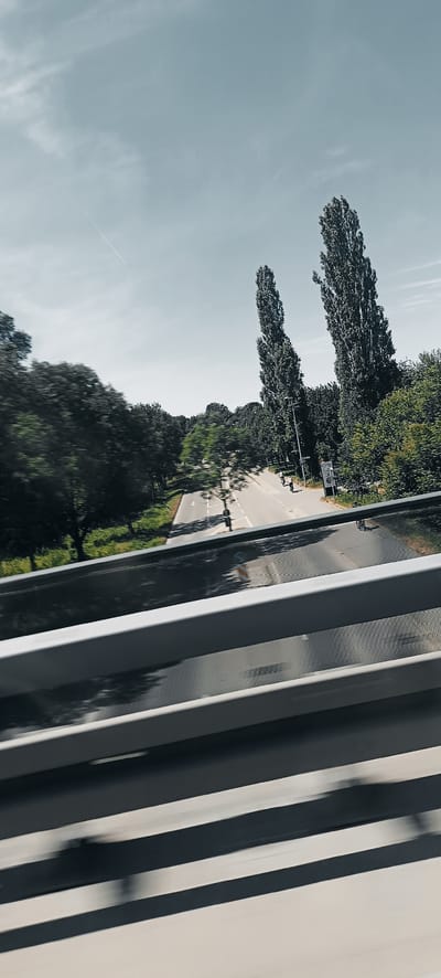 An elevated, oblique, motion-blurred view captures a two-lane road flanked by trees and tall poplar trees. The sky above is a pale blue with a thin contrail from an aircraft. On the right side of the road, a black traffic signpost is visible. Several individuals are seen on bicycles or scooters on the right lane of the road. The foreground is dominated by a grey metal railing, also motion-blurred, with horizontal bars. A black mesh pattern is visible beneath the railing. The scene appears to be from a bridge or overpass looking down onto the road. The location is identified as Waiblingen, Germany.