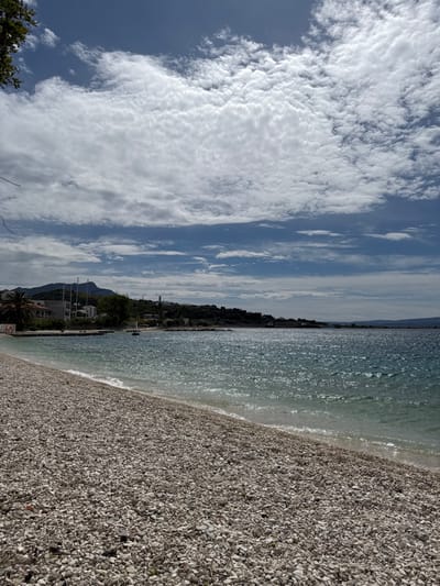 The image shows a view of a beach in Dugi Rat, Croatia, looking towards the sea from the shore. The beach is composed of small, white stones. The water is blue and clear, with small waves lapping at the shore. The shoreline is a mixture of sand and stones. There are trees and buildings in the distance, along the hillside. The sky is blue and cloudy. There are no people or animals in the image.