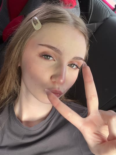 The image is a close-up, selfie-style portrait of a young woman with fair skin and blonde hair. She is wearing a grey t-shirt and has a light-colored hair clip in her hair. Her makeup includes eyeshadow and eyeliner, and her lips are slightly pursed. Her right hand is raised, forming a peace sign. The background appears to be the interior of a vehicle, with a dark, textured seat visible behind her. The lighting suggests it is daytime. There is no indication of the city of Новая Отрадовка, Russia, within the image.