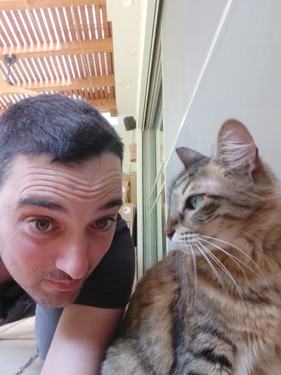 A selfie taken indoors features a man and a cat. The man, fair-skinned with dark hair, is positioned at the top of the frame, his face close to the camera. He is wearing a dark-colored shirt. The cat, a tabby with brown and grey fur, lies closer to the bottom of the frame. Its head is turned upwards, whiskers prominently displayed. The background shows a window with wooden slats and an interior ceiling with track lighting. The photo appears to be taken on a table or other flat surface. This location is in Beer Yaaqov, Israel.