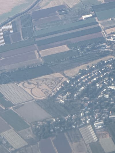 An aerial photograph shows agricultural fields surrounding a residential area in Givat HaShlosha, Israel. The fields are divided into rectangular plots, some with visible irrigation systems. In the center of the image, a large heart shape formed by cleared land or crops is visible within a field. Within the heart, the words "FOR" and "CUCLU" are etched or mown into the land, with a question mark preceding the words. The residential area consists of numerous houses with red tile roofs, interspersed with trees.