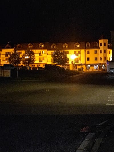 The image depicts a yellow building with multiple stories, illuminated by yellow lights from its windows and streetlights in front. The building has a tower on the right side and several dormer windows on the roof. Trees are visible in front of the building, and cars are parked in the foreground. The scene is at night, with a dark sky. The location is identified as the Hotel Záluží in Nový Jičín, Czechia. The hotel appears to be situated near a road and possibly a park or green space. There are no people visible in the image.