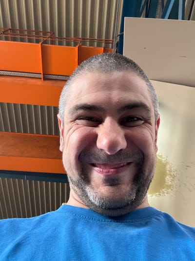 A close-up, selfie-style, medium shot shows a white man from the chest up. He is looking at the camera with a wide smile, his eyes crinkled at the corners. He has short, graying brown hair, a goatee, and a faint gold chain around his neck. He is wearing a blue t-shirt.
The background is out of focus but appears to be an indoor industrial setting. On the left, there are bright orange shelves with a ribbed, silver metal backing. To the right, a large, pale beige panel dominates the background, with a darker mark visible on its surface. A blue metal structure is also visible in the upper right quadrant.
The image does not provide any specific geographical markers to confirm its location in Ellwangen, Germany.