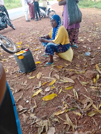 The scene is set in Ugwolawo, Nigeria, on a reddish-brown dirt ground adjacent to a paved road, scattered with dry brown and yellow leaves. In the midground, a woman is seated on a yellow sack acting as a stool. She wears a blue patterned hijab, a yellow long-sleeved top, blue patterned trousers, and light blue flip-flops, holding a small dark object with both hands and looking towards the left. To her immediate left on the ground is a black woven bag containing two yellow plastic jerry cans with red caps. Behind the seated woman, another female figure stands, partially obscured by a large dark bag slung over her shoulder, wearing a light purple skirt and a patterned underskirt. In the background, on and near the paved road, several men are present; one is in white attire, another in maroon trousers, and a third in dark clothing. Two motorcycles are visible: one partially in the left foreground and another further back near the men. A white scooter or moped is also parked near the road. Green foliage borders the road in the distance.