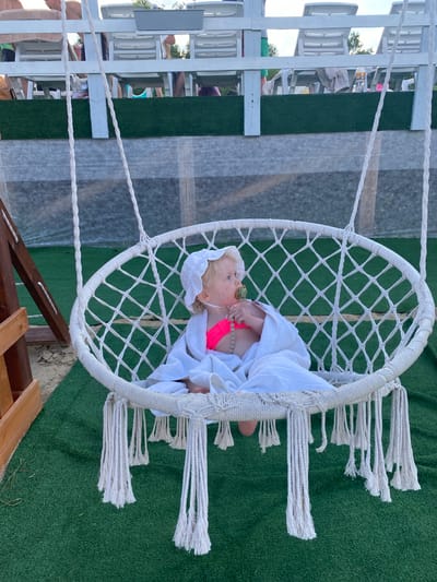 A young child is sitting in a white macrame hanging chair outdoors. The child, wearing a white bonnet and a neon pink swimsuit, is wrapped in a white towel. They are holding a pacifier with a beaded string. The chair is suspended by thick white ropes and has decorative tassels hanging from its bottom edge. The chair sits on a patch of artificial green turf, with sand visible to the left of the frame. In the background, a white wooden structure with rows of empty white lounge chairs is visible, suggesting an outdoor seating area, possibly a resort or beachside location. The sky is overcast. The location is identified as Колокша, Russia, though this information is not visually verifiable from the image alone.