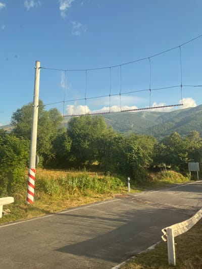 The image depicts a road scene in Makotsevo, Bulgaria, on a clear day with a blue sky and scattered white clouds. A paved road curves to the right, bordered by a white guardrail on the right side and grassy verges with vegetation on both sides.

In the foreground, a red and white striped pole stands vertically on the left side of the road. Extending from this pole is a horizontal wire structure with several vertical wires suspended from it, creating a barrier of sorts across the road. Attached to this structure is a red and white striped bar.

Beyond the road, dense green trees and bushes line the landscape, with a field of dry grass and vegetation to the left of the road. In the background, a range of forested mountains rises under the bright sky. The lighting suggests it is daytime, possibly late afternoon given the shadows. No people are visible in the image.