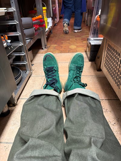 The image depicts a first-person perspective from an individual seated or lying on the floor in what appears to be a commercial kitchen in La Salle-les-Alpes, France. The foreground shows the person's legs, clad in cuffed dark olive-green pants and worn vibrant green suede high-top sneakers with dark laces and light-colored soles. Their feet are pointed towards the background.
The environment is characterized by a tiled floor featuring a repeating octagonal or hexagonal pattern in shades of orange-brown and beige. To the immediate left, a multi-tiered stainless steel utility rack holds various kitchen items, including dark pots or bowls and plastic containers with red and yellow accents. An electrical cord is visible on a lower shelf. To the immediate right, a beige-colored unit with a gridded vent on its lower half stands beside a clear plastic dispenser containing a light-colored liquid.
In the background, a second individual is visible from the knees down, walking away from the camera. This person wears blue denim jeans and light-colored athletic shoes with dark soles. They are traversing a narrow aisle lined with additional stainless steel surfaces and equipment, receding further into the kitchen area. The overall scene suggests an active, working commercial food preparation space.
