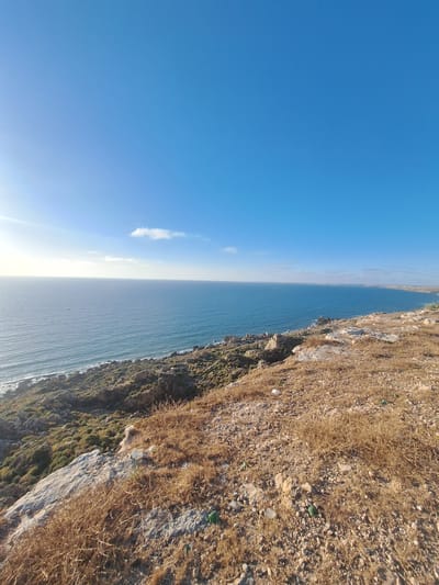 This media file depicts a scenic coastal view, likely taken in Ahrara, Morocco. The image is oriented vertically, showcasing a vast expanse of bright blue sky on the left, with a single wispy white cloud. On the right, a rugged cliff face slopes down towards the sea. The cliff is covered in dry, golden-brown grasses and rocks, with patches of green vegetation clinging to its steeper sections. The sea stretches out horizontally across the mid-ground, appearing calm and blue, with gentle waves lapping at the base of the cliff. The sun appears to be low in the sky, casting a warm light and creating a bright glare on the horizon where the sea meets the sky. The overall impression is one of a serene, natural landscape on a clear day. There are no people or man-made objects clearly visible in the scene, beyond what might be implied by the general setting. The time of day appears to be either early morning or late afternoon, given the angle of the light.
