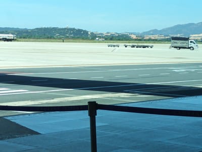 The image depicts an outdoor scene at an airport, likely within the vicinity of Bắc Cam Ranh, Vietnam, given the visible text "CAMRANH INTERN" on a dark barrier in the foreground. The foreground is dominated by a blue tiled area and a dark barrier with white text. Beyond this, a tarmac area is visible with various markings and shadows. In the mid-ground, several airport utility vehicles and equipment are present. To the left, a large white tanker truck with "Skypec" branding is parked. Further to the right, two sets of baggage carts or dollies are positioned on the tarmac, with another white truck with a covered bed visible in the far right mid-ground. A lone individual in a blue shirt is walking on the tarmac in the mid-ground, between the tanker truck and the baggage carts. The background features a landscape of rolling, tree-covered hills with some structures visible on the summits. The sky is clear and blue, suggesting a daytime setting. The overall impression is that of an active airport environment with ongoing ground operations.