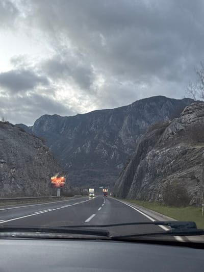 A road winds through a mountainous landscape under a cloudy sky. A large, gray mountain looms to the left, its rocky texture visible. The road, marked with white lines, leads towards two indistinct vehicles in the distance. Closer to the viewer, along the road, stand structures with glowing, possibly illuminated displays. The view is captured from inside a vehicle, with a portion of the dashboard or side visible on the right.