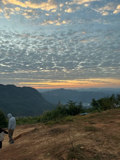 A landscape view shows a sky filled with textured clouds. The horizon line displays a gradient of color, suggesting either sunrise or sunset. Lush green mountains form the backdrop. On the right, a grassy slope with a hint of a dirt path is visible. Two individuals stand on the slope, one with a white hoodie. The image is rotated 90 degrees.