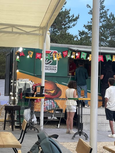This outdoor scene in Daranak, Armenia, depicts a temporary food service setup under a white canopy structure. A dark green food truck, branded "HUMANYAN SHAURMA since 1998" and featuring a large graphic of a hamburger and french fries, serves as the central point. The truck's service window is open, and its exterior is decorated with strung lightbulbs and colorful triangular pennant flags, some bearing "SINCE 90s" text.

Approximately five individuals are visible. Inside the truck, a person in a red shirt is seen. Outside, a man in a dark green shirt and jeans stands facing the truck's service window. To his right, another person in dark clothing, possibly with a hood, also faces the truck. A woman in a white lace top and white shorts stands near a tall, dark metal table with a yellow and green top, which holds a black point-of-sale system and a small black object. A silver Coca-Cola mini-fridge is positioned next to this table. Further to the right, a man in a white polo shirt and dark shorts stands with his back partially to the viewer. In the foreground, parts of wooden tables and chairs are visible, along with a dark blue backpack on one of the tables. The ground is a mix of gravel and possibly covered surfaces. In the background, pine trees are visible against a light blue sky. Power cables are present on the ground.