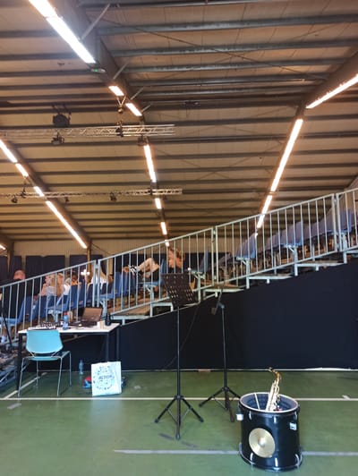 The image depicts an indoor event with a performance or rehearsal taking place. The setting appears to be a large hall with a corrugated metal ceiling and walls, illuminated by long fluorescent lights. The floor is a green turf-like surface, divided by white lines, suggesting it might be a sports hall or a similar venue.
In the foreground, a large black bass drum with a cymbal on its side is set up on a stand, with a saxophone placed inside it. To the left of the drum, a music stand is visible. Behind the drum and music stand, a raised metal structure with seating is occupied by several people, who appear to be musicians. Some are looking down at their instruments or sheet music.
Further in the background, a dark curtain or backdrop is hung, creating a stage area. The overall atmosphere seems to be a focused practice or preparation for a performance, with the participants engaged in their musical activity. The lighting suggests it could be daytime or evening, depending on whether the natural light from outside or the artificial lights are dominant. There is no visible indication of weather conditions, and the emotions conveyed are one of concentration rather than overt joy or excitement.
On a small table on the right, a white bag with text that appears to say "ACTION" and some other symbols is visible, along with a bottle of water. The context of "Warffum, Netherlands" suggests this is a local event in that region.