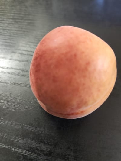 A single ripe apricot rests on a dark, textured surface that appears to be a table. The apricot is plump and round, with a gradient of color from a soft peachy orange to a deeper blush on one side. Small, scattered specks of a darker hue are visible on its surface. The texture of the surface beneath the fruit is characterized by parallel lines, suggesting wood grain or a similar material with a distinct pattern. The lighting in the scene is soft, casting a gentle glow on the apricot and highlighting its smooth skin. There is no text visible in the image, and the focus is solely on the fruit and its immediate surroundings. The image appears to be a close-up, still-life composition.