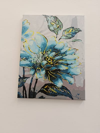 A framed painting of a stylized blue flower with yellow and black accents is displayed on a white, textured wall. The painting features a close-up view of the flower, with its petals rendered in various shades of blue and outlined in yellow. The center of the flower is a complex mix of yellow and black. Several leaves, painted in shades of green, blue, and yellow, surround the flower, with some appearing to droop. The background is a muted gray, with subtle grey and pinkish-grey tones that create a soft, textured effect. The painting has a contemporary, almost abstract quality due to the bold brushstrokes and simplified forms. The overall impression is one of artistic interpretation rather than photorealism. The painting is hung on a wall in what appears to be an indoor setting, likely a home given the plain white wall. There is no visible text in the image.