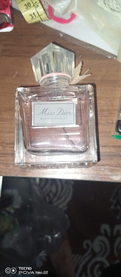 A close-up, eye-level, indoor shot displays a glass bottle of Miss Dior Blooming Bouquet perfume. The bottle is rectangular with a clear cap and a pink band around the neck. The label on the bottle reads "Miss Dior" in cursive and "BLOOMING BOUQUET" in smaller text. The bottle contains a pinkish liquid. The background consists of a wooden surface and other out-of-focus objects. The bottom of the image shows a blurred reflection of the surroundings, including the "TECNO POVA Neo 2" logo. The image is taken in a domestic setting, likely a home, within the city of Tahmach, Uzbekistan.