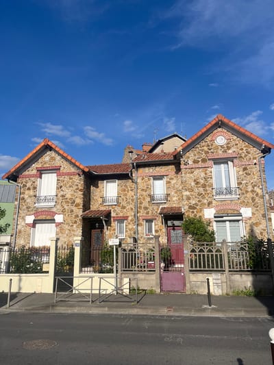 The image depicts a residential street scene in Vitry-sur-Seine, France, under a clear blue sky with scattered white clouds. The setting appears to be a sunny day, likely midday or early afternoon, given the bright illumination.
The main subjects are a row of stone and brick houses, closely situated along a paved street. The houses feature facades constructed from irregular, light-colored stones interspersed with reddish-brown brickwork. The bricks are used to outline windows, create decorative arch patterns above some windows, and form vertical accent strips. The roofs are covered with distinct reddish-orange tiles, and several white-framed windows with white louvered shutters are visible. Some windows also have elaborate black wrought-iron grates.
At street level, various metal fences and gates separate the properties from the sidewalk. One prominent house entrance features a dark red or maroon wooden door with decorative ironwork, beside which is a matching maroon metal gate. Another section of fencing is made of concrete or stone with decorative cut-outs. Green foliage and shrubs are visible behind some of the fences. The asphalt street runs parallel to the houses, featuring a manhole cover and a black bollard on the adjacent concrete sidewalk.
Notable text includes a white sign on a pole that reads "vitry en scene" and "point info" with a red logo. Further down the street, a house number "24B" is visible on a blue plaque attached to a white pillar. Another partially obscured street sign appears to indicate "Rue Gabriel Peri," which confirms the location. No people or active events are depicted in the static scene.