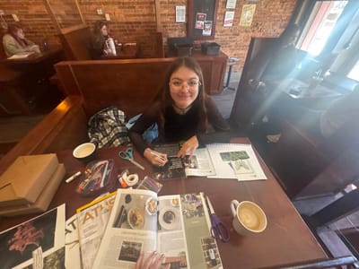A young woman with glasses sits at a booth table, surrounded by art supplies. Magazines, scissors, glue, and a pencil case are scattered around her. She appears to be working on a collage, with cut-out images and sketches visible on a sketchbook. A mug of coffee sits nearby. Other patrons occupy booths in the background of the cafe-like setting. The interior has brick walls and dark wood booths.