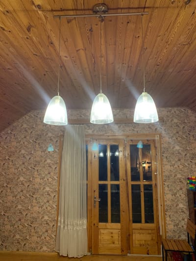 The image shows an interior scene, likely a room in a house, featuring a wooden-paneled ceiling with three hanging pendant lights. Each light has a clear, bell-shaped glass shade. Below the lights, there is a double door made of wood with glass panes. To the left of the door, a sheer, light-colored curtain hangs. The walls are covered in a textured wallpaper with a pattern of oval shapes in various shades of brown and beige. The floor appears to be wooden. In the background, to the right of the door, a shelf unit with colorful blocks is partially visible. The scene is well-lit, primarily by the pendant lights. No people are visible in the image.