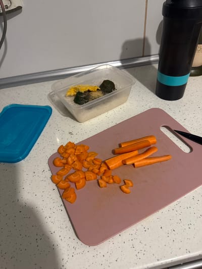 On a countertop with a speckled white surface, a pink cutting board holds partially sliced carrots. A container to the left contains food items, and a blue lid sits next to it. A black object with a blue band is positioned in the upper left corner.