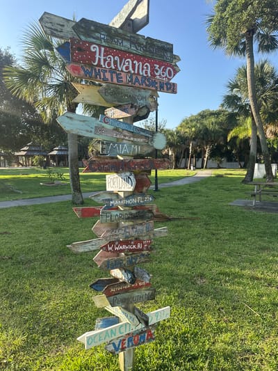 This media file shows a directional signpost with numerous wooden signs pointing in various directions, indicating distances to different cities and locations. The signs are weathered and hand-painted, with some text legible. Visible destinations include Havana, White Marsh MD, Boardman OH, Chicago IL, Miami, Portland, Marathon FL, Florida, W. Warwick RI, Metuchen N.J., Plymouth MA, and Ocean City. The signpost is set in a grassy area with palm trees and other lush greenery in the background, suggesting a warm, sunny climate. The lighting indicates it is daytime, likely morning or afternoon, with clear blue skies. The overall scene evokes a sense of travel, exploration, and perhaps a bit of whimsy. The location is identified as Melbourne Beach, United States of America. There are no people visible in the image.