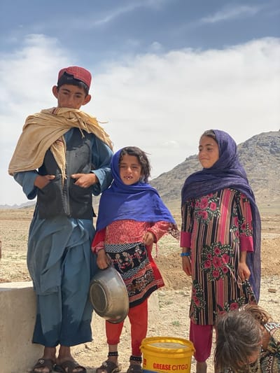 Three children stand near a water source in a rural, arid environment, likely in or around Ashoqah, Afghanistan. The tallest child, a boy, wears a red cap, a light-colored shawl draped over his shoulders, a dark vest over a blue tunic, and matching blue pants. He stands to the left, resting his hand inside his vest. A younger girl in the center wears a blue head covering, a red top over pants, holds a metal pot. A second girl on the right wears a blue head covering and a long floral-patterned dress over pink pants. A yellow bucket with "GREASE CITGO" printed on it sits on the ground in front of them. The background shows a sparse landscape with a mountain range in the distance and a cloudy sky. The ground is dry and rocky.