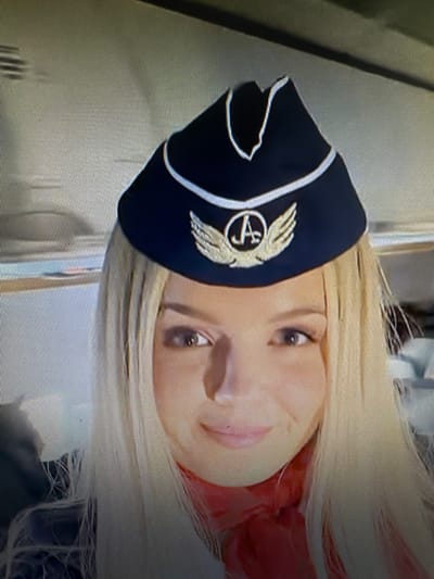 The image shows a close-up, rotated portrait of a woman wearing a dark blue pilot's hat with a gold emblem. The hat has white trim. The woman has blonde hair and is looking towards the camera with her mouth slightly open. Her eyes are a light brown color. She is wearing a red scarf or similar garment around her neck. The background appears to be indoors, possibly on an airplane or in a similar setting, with muted, neutral-colored surfaces. The lighting suggests it is daytime. There is no visible text in the image.