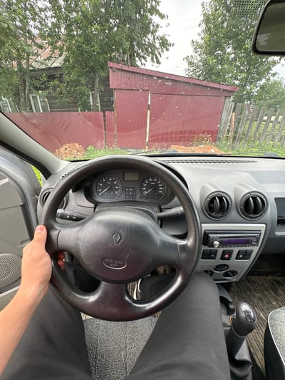 The image shows the interior of a car, seen from the driver's perspective. A person's left hand is on the steering wheel, which has a Renault logo in the center. The dashboard features a speedometer and tachometer. A car stereo is visible, and the gear shift is in the center console. Outside the car, a red corrugated metal building and a wooden fence are visible, along with trees and vegetation. The scene appears to be in a rural or semi-rural setting, possibly in Yayva, Russia, given the architectural style of the building and the fence. The sky is overcast, and there are water droplets on the windshield.