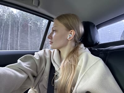 A young woman with blonde hair pulled into a ponytail is sitting in the back seat of a car, looking out the window. She is wearing a light-colored hooded sweatshirt and has white wireless earbuds in her ears. The car appears to be moving through a wooded area, as seen through the window. The image was captured in Didino, Russia.