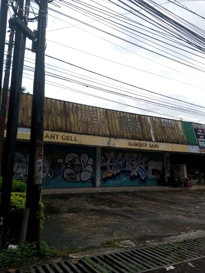 The scene depicts a street view of a commercial building in the city of Kamji Na-Yekha, Bhutan. Overhead, numerous black electrical or communication wires span across a light grey, overcast sky. Two dark, weathered utility poles stand in the left foreground. The commercial building features a faded yellow-gold corrugated metal upper facade, showing signs of wear and dark staining. Below this, a horizontal band of lighter yellow-beige spans the building. Two central storefronts are closed and covered extensively with multi-colored graffiti, including a white and black cartoonish design, stylized lettering such as "DEBAT," "SLEP," "YAMOR," and "AMASKLY." Above these graffiti-covered sections, partial signs read "ANT CELL" and "SUMBER SARI" in dark lettering. To the right, an adjacent storefront appears open, displaying stacked white sacks and a red object; a black scooter or motorcycle is parked in front. Text on this section of the facade includes "TOKO KARTIK & BARU" and "TYBO." The foreground features dark, wet-looking asphalt or concrete pavement, with a dark metal drainage grate running along the bottom edge. Green foliage and weeds grow near the utility poles and the pavement's edge on the left. No individuals are visible.