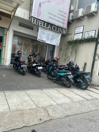 The image shows a street scene in front of a business establishment in Bojongsoang, Indonesia, located at the coordinates -6.972, 107.636. The business is called "Luella Clinio" and advertises skincare, slimming, eyelash extension, and brow embroidery services. Five motorcycles are parked in front of the business, all facing the street. The motorcycles are black, and the front two are a lighter blue. Some motorcycles have license plates visible. The street is paved with concrete, and there is a sidewalk along the front of the building. There is a green vine growing up the wall of the building, and a small amount of greenery in the background. The day is overcast.